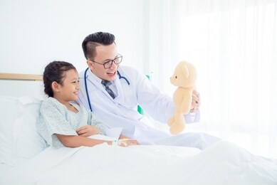 pediatrician male doctor holding teddy bear and playing with child patient who lying on hospital bed