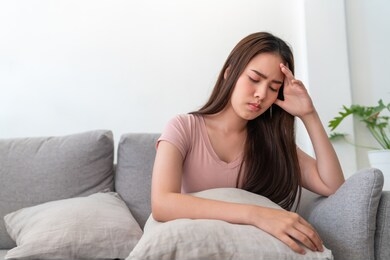 asian young woman holding her head,she has a headache sitting on sofa couch in living room at home.