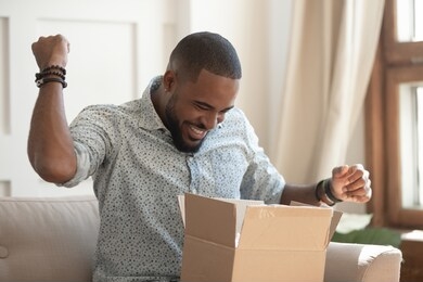 excited african man customer receive good parcel open cardboard box at home satisfied with great purchase, happy black male consumer unpack package look inside overjoyed by postal shipping delivery