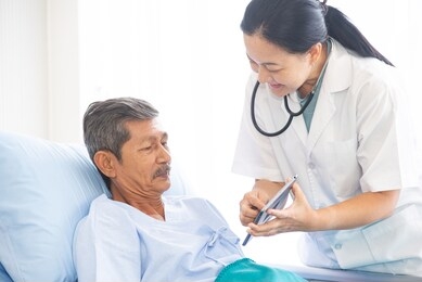 asian woman professional doctor with clipboard visiting and diagnosing the old man patient lying in bed at hospital ward. physician has a routine work to check up and have consulation the patient