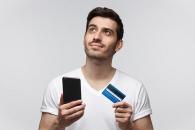 young handsome man thinking about online shopping via internet, holding credit card and smartphone isolated on white background