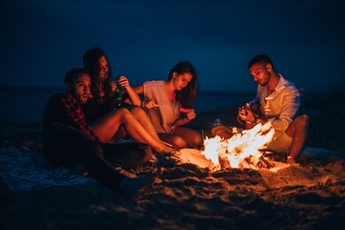 young couple enjoying with friends around campfire on the beach at night by the sea while listening to a guitar songs