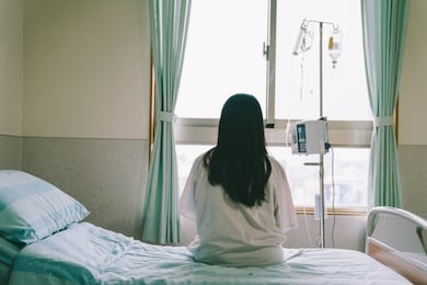 back view of asian chinese female patient with dropper looks out the window of hospital room. healthcare concept. lonely woman with long black hair sitting on bed wants to go outside enjoy sunshine