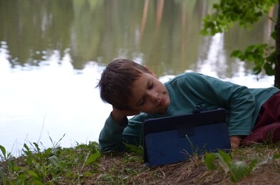 boy schoolboy with a tablet in a beautiful meadow near the lake plays games, watches cartoons. boy child playing with tablet pc outdoor with forest on background computer game dependence concept