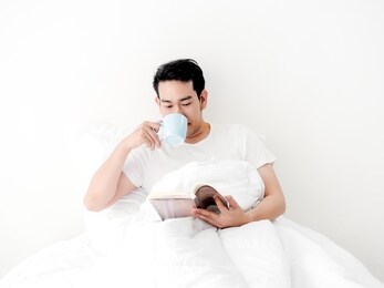 happy asian man reading a book and drinking coffee on bed, man lifestyle on weekend concept.