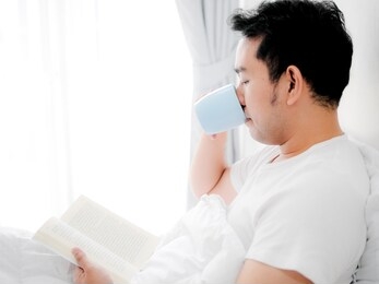 happy asian man reading a book and drinking coffee on bed, man lifestyle on weekend concept.