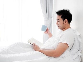 happy asian man reading a book and drinking coffee on bed, man lifestyle on weekend concept.