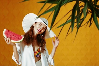 happy smiling fashionable woman wearing white wide brim hat holding watermelon slice. yellow background. copy, empty space for text