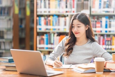 asian young student in casual suit doing homework and using technology laptop in library of university or colleage with various book and stationary over the book shelf background, back to school