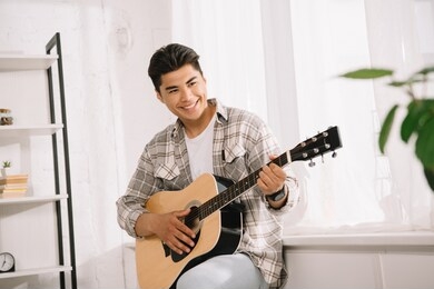 handsome, young asian man looking away while playing acoustic guitar at home