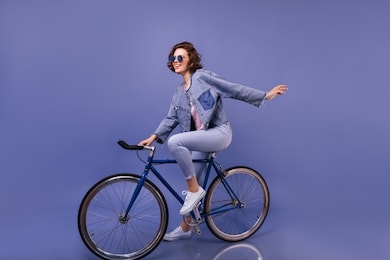 amazing woman in spring clothes sitting on bicycle. indoor portrait of lovely girl in sunglasses fooling around on violet background.