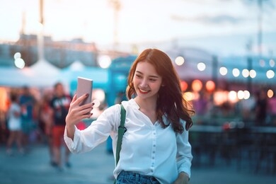 happy young travel asian woman using mobile phone and selfie on street market against light bokeh background at dusk in bangkok, thailand, travel vacation city concept