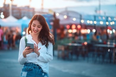 happy young travel asian woman using mobile phone and relax on street market against light bokeh background at dusk in bangkok, thailand, travel vacation city concept