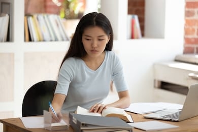 concentrated young asian female student sitting at desk in library. focused millennial girl studying, preparing for entrance exams or university tests, reading book, taking down notes in textbook.