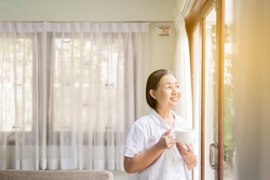 senior asian woman holding a cup of coffee near the window in the morning,happy and smiling,positive thinking,health insurance concept