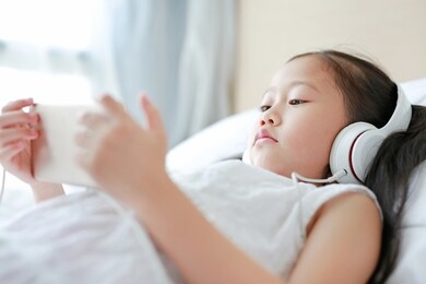 portrait little asian girl using headphones listen music by smartphone while lying on bed at home.