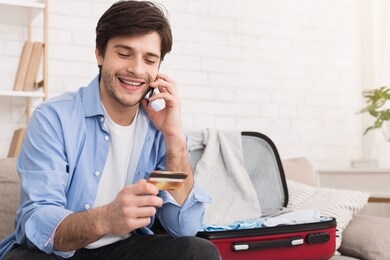 man booking hotel, talking on phone and preparing for vacation with open suitcase nearby