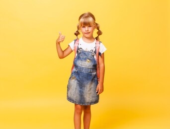 happy little girl with blond hair and with pink backpack jumps in jeans overalls on a yellow background. jeans denim. 