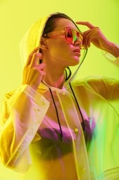 fashion portrait of an attractive young asian woman with long wet hair standing isolated over yellow background, posing in transparent raincoat and sunglasses
