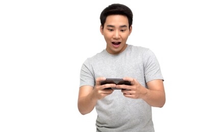 portrait of an excited asian man in t-shirt playing games on mobile phone isolated over white background