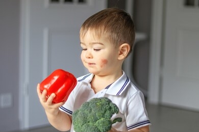 little boy with atopic dermatitis on his cheek holding sweet red pepper and fresh broccoli in his hands looking at it hesitate to eat. food allergy. child eczema. 