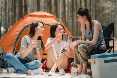 group of young asian friends camping or picnic together in forest, teenager female enjoy moment talking in front of their tent. women do adventure activity and travel on holidays vacation in summer.