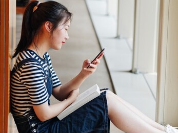 young student girl with ponytail sitting on floor in library and leaning on shelf while using her smart phone with book on her legs.