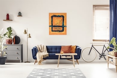 yellow and blue painting hanging on white wall in bright living room interior with grey cupboard, gold lamp, sofa with blanket and pillows, and bike standing under window with blinds