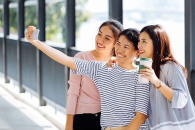 young asian people taking selfie photos together inside the glass building. three beautiful women having fun taking pictures outdoors in blurred background