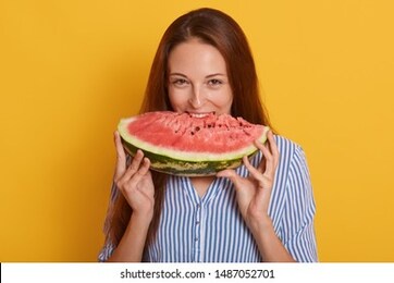 close up portrait of young woman biting slice of watermelon while looking directly at camera, posing isolated over yellow background, adorable female wearing striped shirt. healthy eating concept.