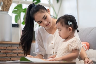 asian pretty young mother and her daughter reading tale story book together. the mom looking the little girl  with happiness. good relationship of mom and kid. family activity and education concept.