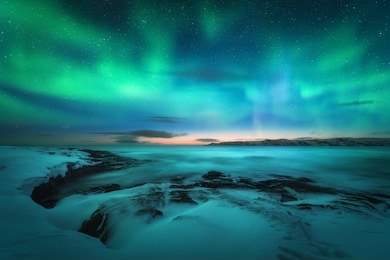 aurora borealis over rocky beach and ocean. northern lights in teriberka, russia. starry sky with polar lights. night winter landscape with aurora, sea with stones in blurred water, snowy mountains