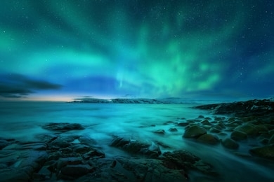 aurora borealis over rocky beach and ocean. northern lights in teriberka, russia. starry sky with polar lights. night winter landscape with aurora, sea with stones in blurred water, snowy mountains