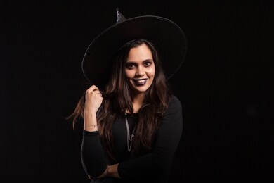 young beautiful witch with a black dress and a big hat smiling over black background. pretty girl ready for halloween dressed up like a witch.