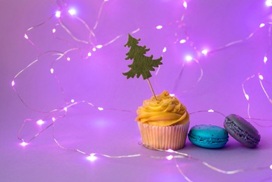 fresh yellow cupcake with the christmas tree and the garland around. bright macaroons near. celebrative creative composition, trend neon light. holiday concept, selective focus, copy space