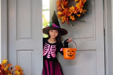kids trick or treat on halloween. little asian girl at decorated house door with autumn leaf wreath and pumpkin lantern. child in witch and skeleton costume and hat with candy bucket. fall decoration.
