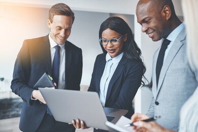 group of diverse businesspeople smiling while standing in a bright modern office discussing work together over a laptop