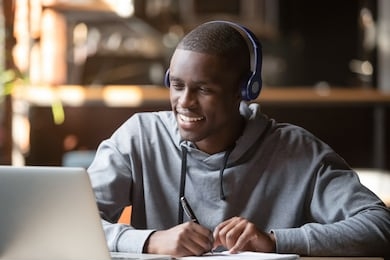 smiling african young man student wear headphones looking at laptop screen writing notes, happy black guy e-learning in internet study online with teacher on computer watch webinar sit in cafe