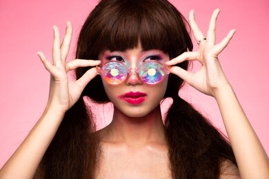 fashion model looks at camera for shooting new collection sunglasses.  beautiful asian woman set afro hair trend make up wear kaleidoscope glasses with colorful, studio lighting pink background