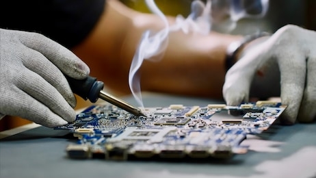 technician engineer in workshop. repairman in gloves is soldering circuit board of electronic device on the table, hands close up. he takes tin with a soldering iron and puts it on microcircuit.
