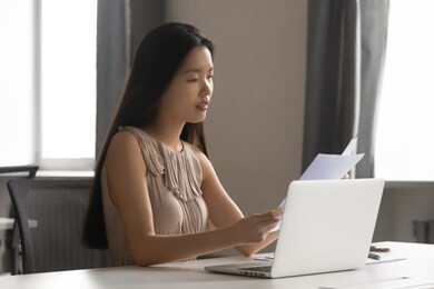 serious focused busy young female asian employee sitting at workplace with computer, holding documents in hands, reading financial paper report, checking bank loan or client invoice, doing paperwork.