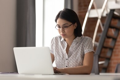 serious focused asian female employee wearing glasses, working with computer in modern loft office. concentrated business woman corresponding with corporate client, checking financial documentation.