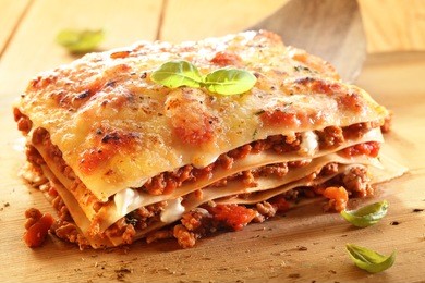 golden lasagne with meat, tomatoes, cheese sauce and pasta in alternating layers on a wooden board garnished with basil
