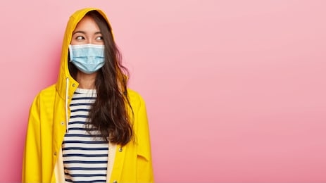 photo of brunette young ill woman wears protective medical mask, has infectional disease, dressed in yellow raincoat, covers head with hood, looks aside, isolated on pink background, copy space area