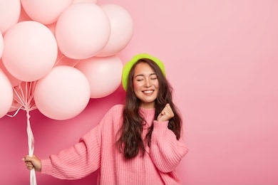 photo of happy pleased brunette asian lady stands with balloons, enjoys cool party with friends, wears beret and loose jumper, celebrates anniversary, poses against pink background. festive event