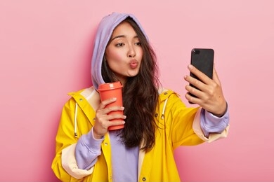 lovely teenage girl with asian appearance, keeps lips folded, sends kiss at camera of cellphone, takes selfie, enjoys drink, wears sweatshirt, hood on head, yellow raincoat, has walk after rain