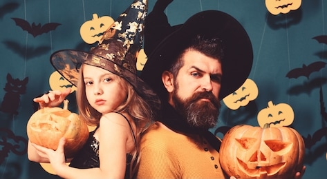 father and daughter in costumes. girl and bearded man with proud faces on green grey background with decor. halloween party and celebration concept. wizard and little witch in black hats hold pumpkins
