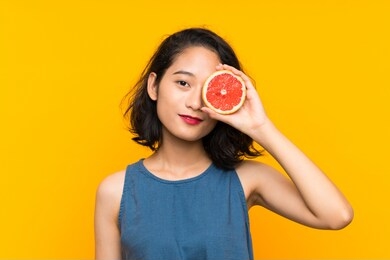 young asian girl holding a grapefruit over isolated orange background