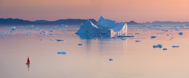 little red sailboat cruising among floating icebergs in disko bay glacier during midnight sun season of polar summer. ilulissat, greenland. unesco world heritage