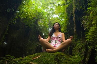beautiful dreamy portrait of young attractive asian woman enjoying nature sitting lotus pose doing yoga meditating relaxed at fresh and ethereal green forest feeling magical and enchanted
